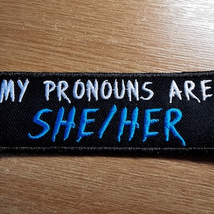 May include: Black fabric patch with white text that reads "My pronouns are" and blue text that reads "she/her".