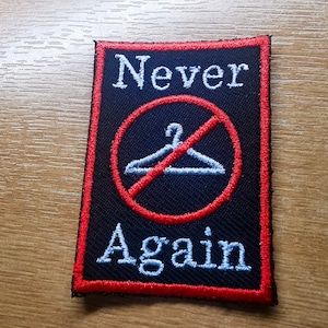 Puede incluir: Parche bordado negro con borde rojo. El parche presenta un círculo rojo con una línea diagonal que lo atraviesa y una percha blanca en su interior. El texto "Never" está por encima del círculo y "Again" por debajo.