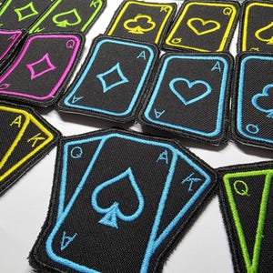 May include: Embroidered playing card patches in various suits and colours. The patches feature black backgrounds with neon green, yellow, pink, and blue stitching. The cards include the Ace, King, and Queen.