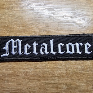 Puede incluir: Parche de tela negro con texto bordado en blanco que dice "Metalcore" en una fuente gótica.