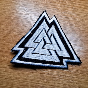 Könnte beinhalten: Ein gestickter Patch in Schwarz und Weiß mit einem dreieckigen Design. Das Design ist ein dreifach ineinandergreifendes Dreieck, auch bekannt als Valknut-Symbol.