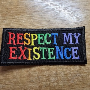 Op de afbeelding: Een zwarte geborduurde patch met de woorden "Respect My Existence" in regenboogkleuren.
