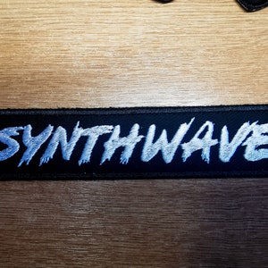 Puede incluir: Un parche de tela negro con texto bordado en blanco que dice "SYNTHWAVE".