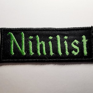 Op de afbeelding: Zwarte stoffen patch met groene geborduurde tekst die "Nihilist" luidt.
