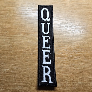 Könnte beinhalten: Schwarzer Stoff-Patch mit weißen gestickten Buchstaben, die vertikal "QUEER" buchstabieren.