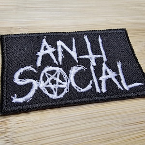 Puede incluir: Parche rectangular negro con texto bordado blanco «ANTI SOCIAL» y un símbolo de pentagrama. El parche está sobre una superficie de madera.