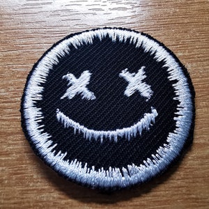 Op de afbeelding: Een geborduurde patch in zwart en wit met een smiley-gezicht met X'en voor de ogen.