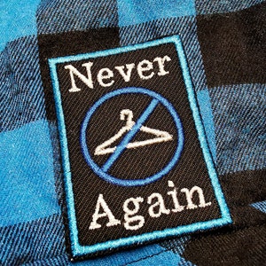 Puede incluir: Un parche bordado negro y azul con el texto "Never Again" y un círculo azul con una percha blanca tachada.