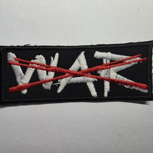 Könnte beinhalten: Schwarzer rechteckiger Aufnäher mit dem Wort "WAR" in Weiß, durchgestrichen mit zwei roten Linien. Der Aufnäher hat einen schwarzen Rand und ist zum Aufnähen oder Aufbügeln auf Kleidung oder Accessoires gedacht.