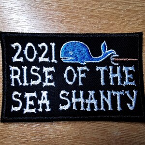 May include: A black embroidered patch with white text that reads "2021 Rise of the Sea Shanty". A blue whale with a hook is embroidered above the text.