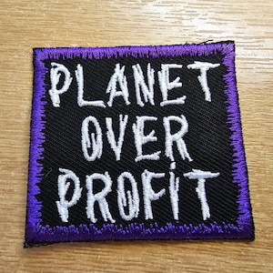 Könnte beinhalten: Ein schwarzer gestickter Aufnäher mit lila Nähten. Der Aufnäher trägt die Aufschrift "Planet Over Profit" in weißen Buchstaben.