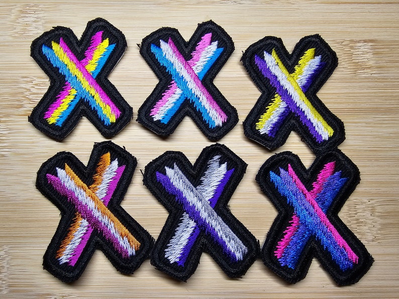 May include: Six embroidered iron-on patches in the shape of an 'X' with different color combinations. The patches are black with a white outline and feature stripes of pink, blue, yellow, white, orange, and purple.