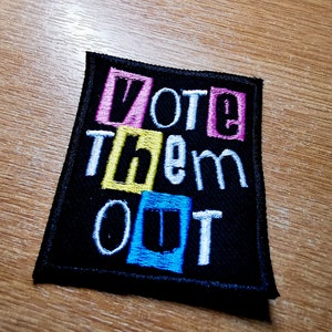 Może przedstawiać: Czarna łatka z tkaniny z białym haftem, na której widnieje napis "Vote Them Out". Słowa znajdują się w kolorowych ramkach: różowej, żółtej i niebieskiej.