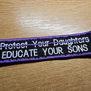 May include: A rectangular black patch with white embroidered text that reads "Protect Your Daughters EDUCATE YOUR SONS." The patch has a purple border and is set against a wooden surface.