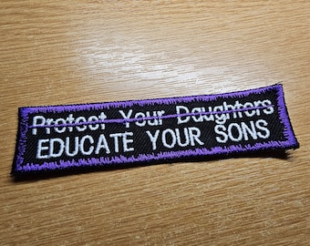 以下が含まれることがあります： 「Protect Your Daughters EDUCATE YOUR SONS」と白い刺繍が施された長方形の黒いパッチ。 パッチは紫色の縁取りがあり、木製の表面に置かれています。