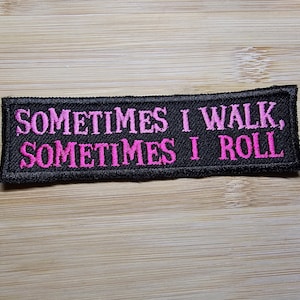 Puede incluir: Parche bordado negro con el texto "SOMETIMES I WALK, SOMETIMES I ROLL" en letras rosa brillante. El parche es rectangular y tiene un borde negro. El fondo es una superficie de madera marr&oacute;n claro.
