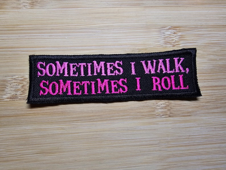 Puede incluir: Parche bordado negro con el texto "SOMETIMES I WALK, SOMETIMES I ROLL" en letras rosa brillante. El parche es rectangular y tiene un borde negro. El fondo es una superficie de madera marr&oacute;n claro.