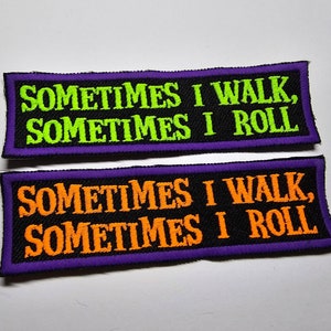 Op de afbeelding: Twee geborduurde patches met paarse randen. De bovenste patch heeft een zwarte achtergrond met de tekst "SOMETIMES I WALK, SOMETIMES I ROLL" in groen. De onderste patch heeft een zwarte achtergrond met de tekst "SOMETIMES I WALK, SOMETIMES I ROLL" in oranje.
