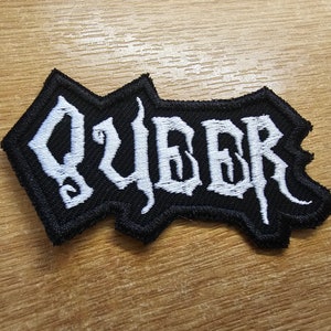 Könnte beinhalten: Schwarzer gestickter Aufnäher mit dem Wort "QUEER" in weißen Buchstaben.