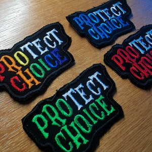 Peut inclure: Quatre patchs en tissu noir avec le texte "Protect Choice" en broderie de différentes couleurs. Les couleurs sont arc-en-ciel, bleu, rouge et vert.