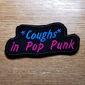 May include: Black embroidered patch with the text "*Coughs* in Pop Punk" in pink and blue lettering.