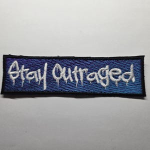 May include: A rectangular patch with the words "Stay Outraged" embroidered in white on a blue background. The text has a dripping effect, and the patch is outlined in black.