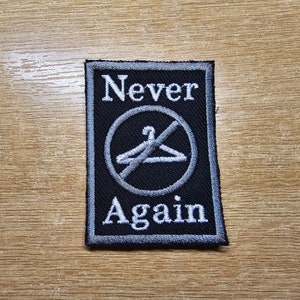 Puede incluir: Parche bordado en blanco y negro con un círculo y una línea que atraviesa una percha. El texto "Never Again" está arriba y abajo del círculo.