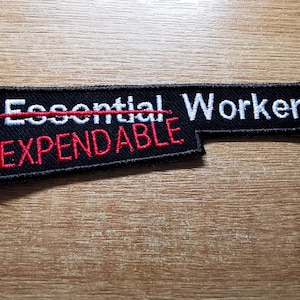 Könnte beinhalten: Schwarzer Patch mit weißem Text, der "Essential Worker" lautet, mit einem roten Strich durchgestrichen. Unter der Linie steht roter Text, der "Expendable" lautet.