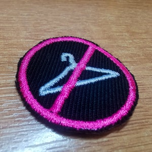 Puede incluir: Un parche bordado negro con un círculo rosa alrededor. El círculo tiene una línea diagonal que lo atraviesa y una percha blanca en su interior. El parche es una señal de prohibido colgar.