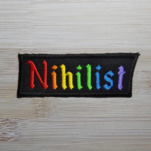 Op de afbeelding: Een zwarte stoffen patch met het woord "Nihilist" geborduurd in regenboogkleuren.