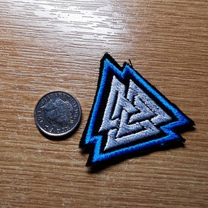 May include: A blue and silver embroidered patch with a triangular design. The patch features a stylized symbol of three interlocking triangles.