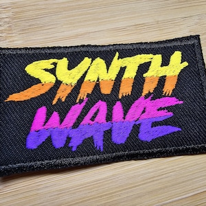 Puede incluir: Un parche de tela negro con las palabras "Synth Wave" bordadas en amarillo, naranja, rosa y morado.