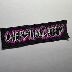 May include: Black rectangular patch with the word "OVERSTIMULATED" embroidered in white, outlined with a pink, spiky design. The patch has a black border and is likely designed to be sewn or ironed onto clothing or accessories.