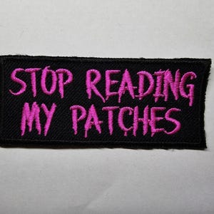 May include: Black embroidered patch with the phrase "STOP READING MY PATCHES" in bright pink lettering. The patch is rectangular and has a black border. The text is in a stylized, slightly gothic font.