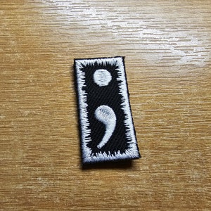 Pode incluir: Remendo de tecido preto com um ponto e vírgula branco bordado e costuras brancas ao redor das bordas.