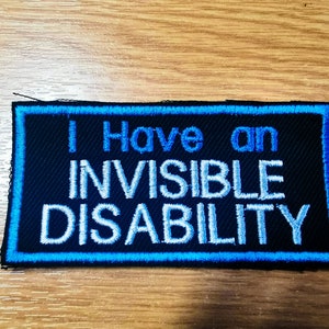 Puede incluir: Parche negro con costuras azules alrededor de los bordes. El texto "I Have an Invisible Disability" está bordado en blanco.