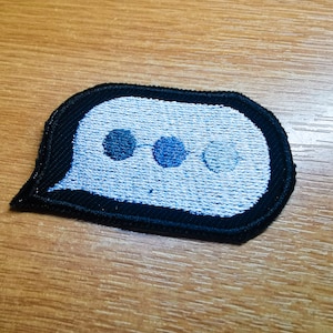 Op de afbeelding: Een geborduurde patch in zwart en wit in de vorm van een spraakballon met drie blauwe stippen erin.