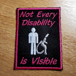 Può includere: Toppa ricamata nera con cuciture rosa. La toppa presenta una silhouette bianca di una persona in piedi accanto a una persona in sedia a rotelle. Il testo "Not Every Disability is Visible" è ricamato in rosa.