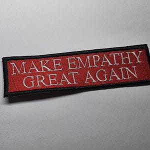 May include: Embroidered patch with the text "MAKE EMPATHY GREAT AGAIN" in white letters on a red background. The patch has a black border and is rectangular in shape.