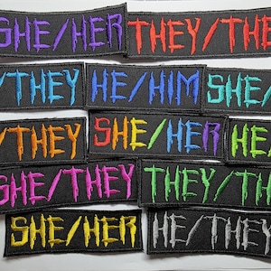 May include: A collection of rectangular black embroidered patches featuring various pronoun combinations in vibrant colors including purple, red, blue, orange, green, pink, yellow, and silver, all rendered in a jagged, distressed-style font.