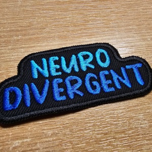Könnte beinhalten: Ein schwarzer gestickter Aufnäher mit dem Text "Neuro Divergent" in blauer Schrift.