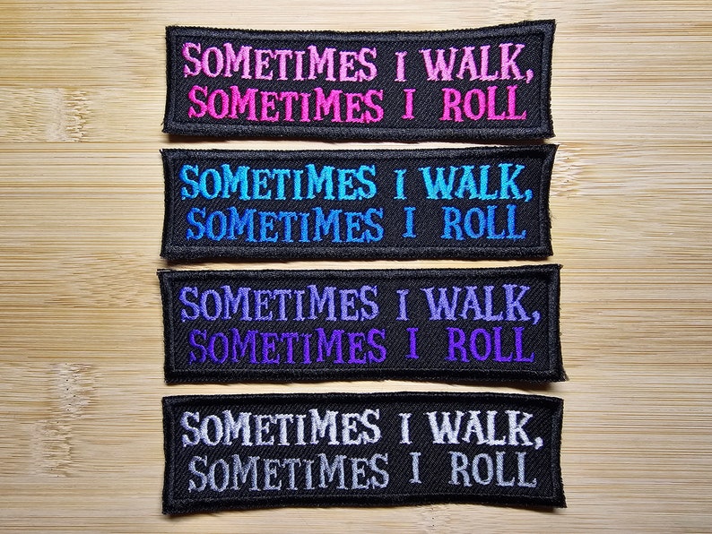 Puede incluir: Cuatro parches bordados con el texto "SOMETIMES I WALK, SOMETIMES I ROLL" en varios colores: rosa, azul, morado y blanco. Cada parche es rectangular con un borde y fondo negros.