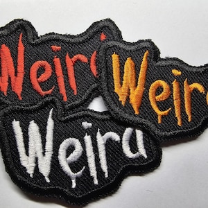 Peut inclure: Trois patchs en tissu noir avec le mot "Weird" brodé en fil rouge, orange et blanc.