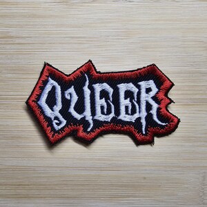 Peut inclure: Patch brod&eacute; rouge et noir avec le mot "QUEER" en lettres blanches.