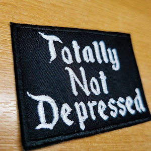 Könnte beinhalten: Schwarzer gestickter Aufnäher mit weißem Text, der "Totally Not Depressed" lautet.