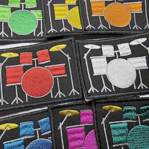 May include: A collection of rectangular embroidered patches featuring a drum kit design on a black background. Each patch displays a drum set in a different color, including green, yellow, orange, red, white, blue, pink, and teal, with yellow cymbals.