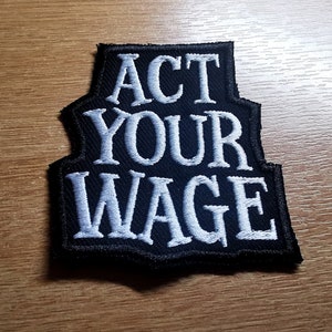 May include: Black and white embroidered patch with the text "ACT YOUR WAGE" in a white font.