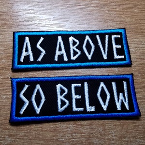 以下が含まれることがあります： 白い文字と青い縁取りのある黒い刺繍パッチが2つ。上のパッチには「AS ABOVE」、下のパッチには「SO BELOW」と書いてあります。