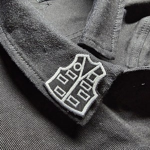 May include: Close-up of a black denim jacket collar with a white and black embroidered patch. The patch features a shield design with geometric shapes. The jacket has visible stitching and buttons.