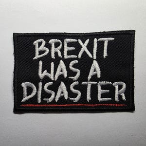 May include: A black rectangular patch with white embroidered text that reads "BREXIT WAS A DISASTER." A thin red line is embroidered beneath the text. The patch is likely designed to be sewn onto clothing or bags.
